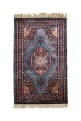 Vista superior completa de una alfombra oriental con fondo gris azulado y medallón central floral en tonos rosado, crema y azul oscuro.