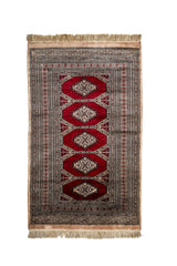 Alfombra oriental vista desde arriba con diseño geométrico central en fondo rojo y bordes con motivos intrincados en tonos beige.