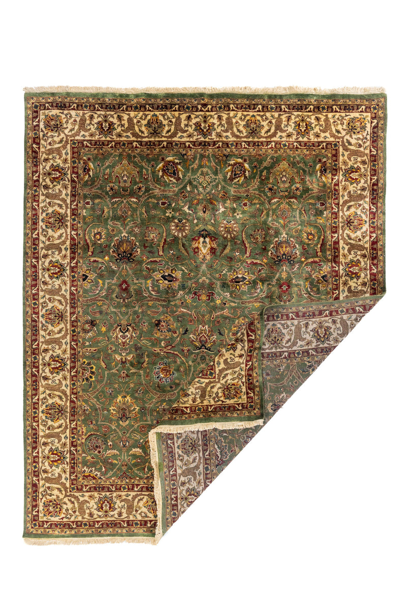 Alfombra verde con borde beige y detalles rojos, esquina doblada mostrando reverso y flecos.