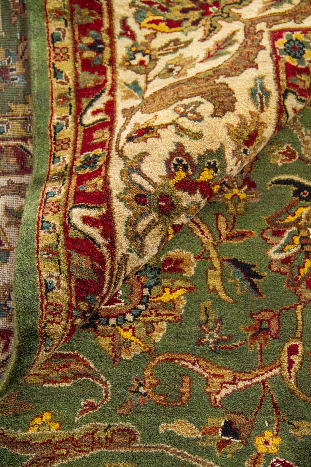 Detalle de pliegue de alfombra verde: textura del tejido, flecos y franja floral multicolor.