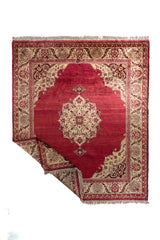 Alfombra roja burdeos con medallón central marfil y borde floral beige, esquina levantada mostrando el reverso.