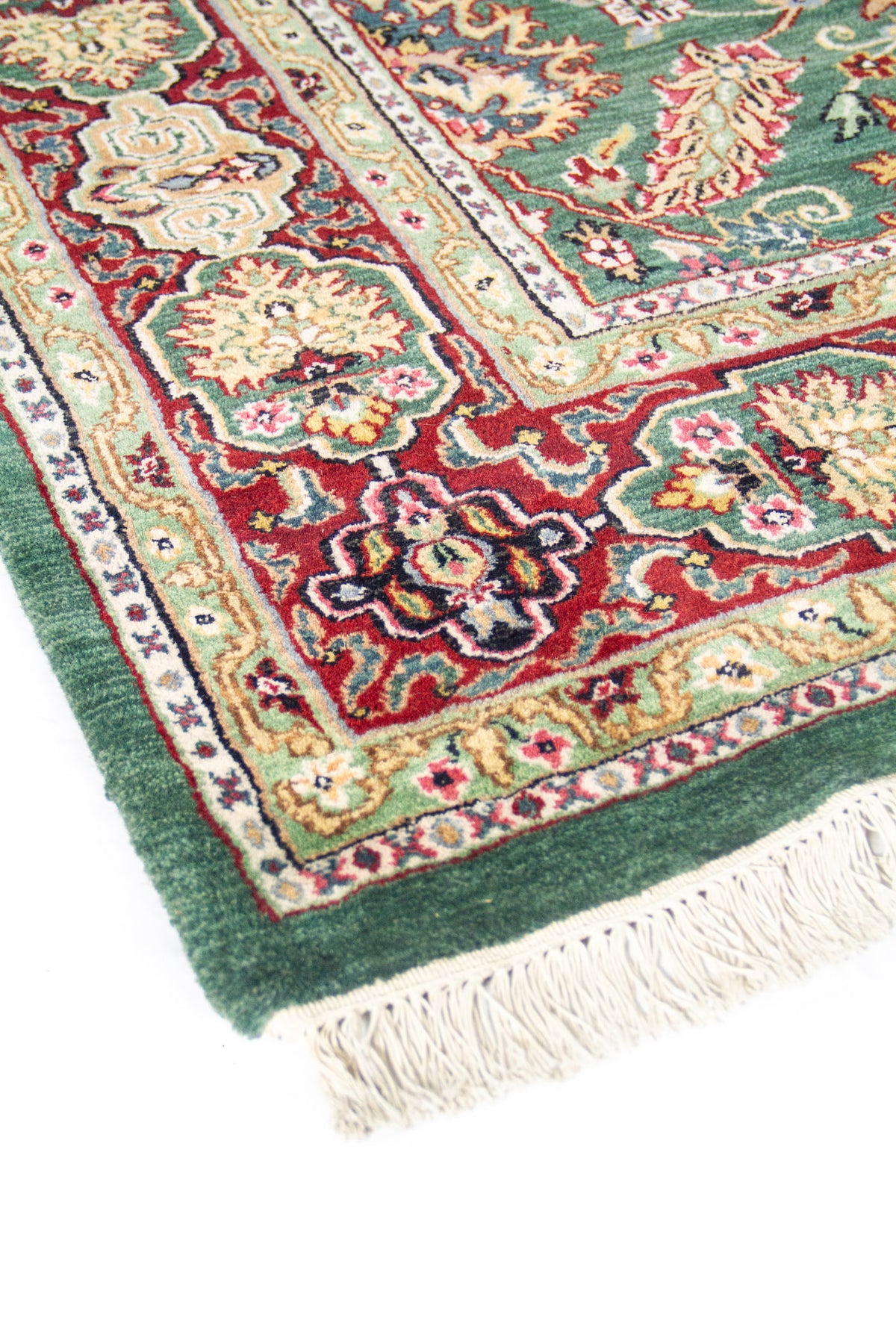 Vista en ángulo de alfombra verde con guarda rojo y beige, mostrando el diseño floral y flecos en un extremo.