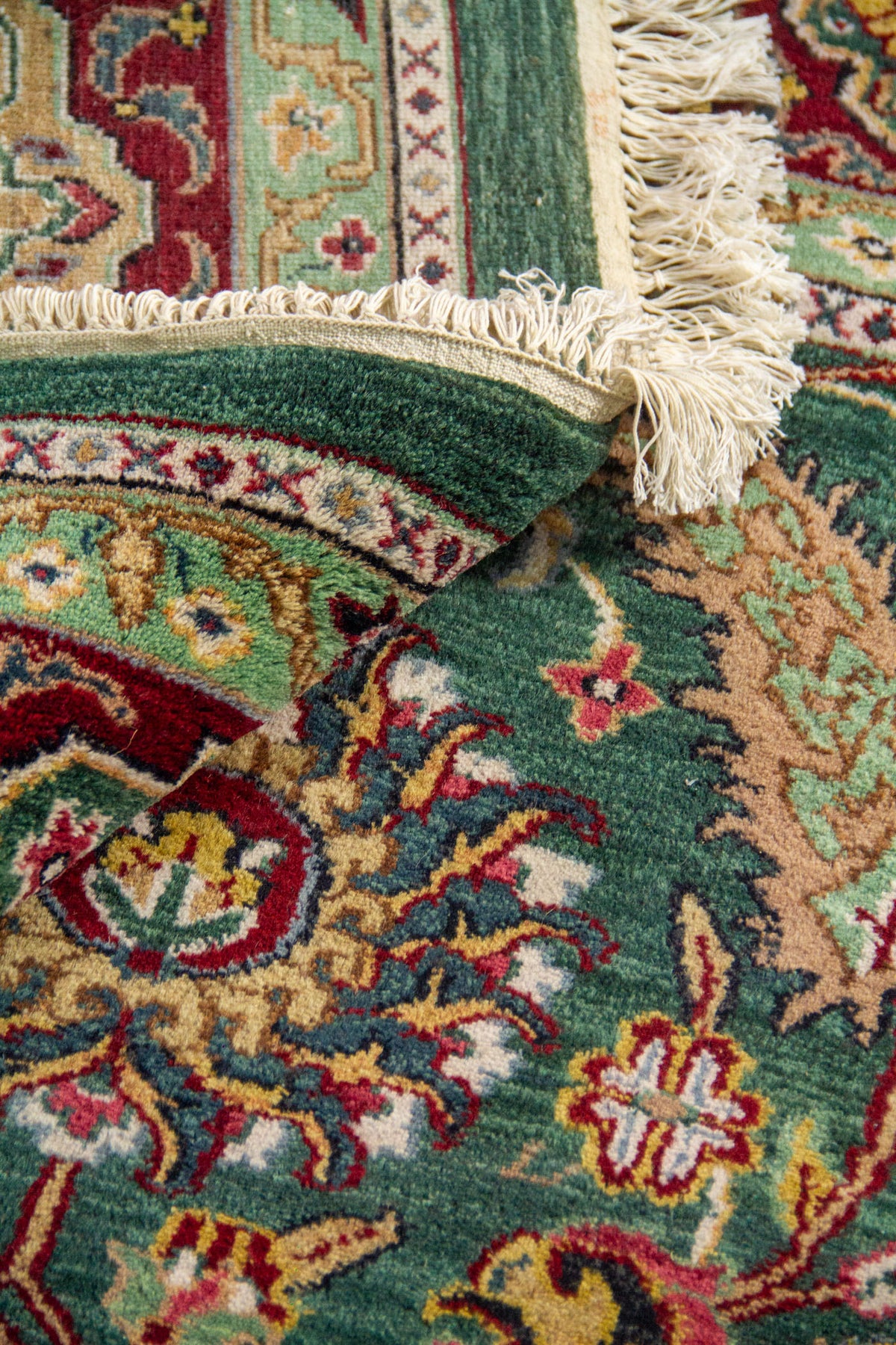 Detalle de alfombra plegada con flecos y textura de pelo corto, mostrando motivos florales sobre fondo verde.