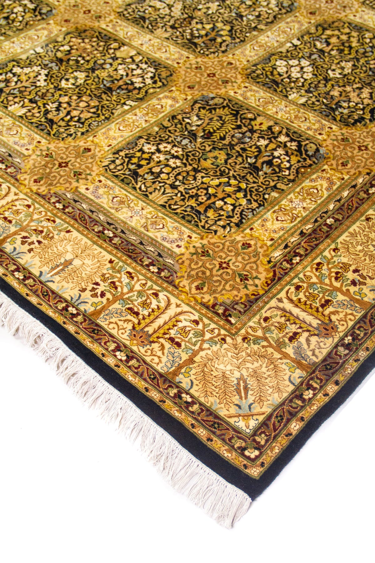 Detalle en esquina de alfombra oriental con flecos color marfil, guarda con motivos vegetales y paneles florales en tonos dorados y negro.