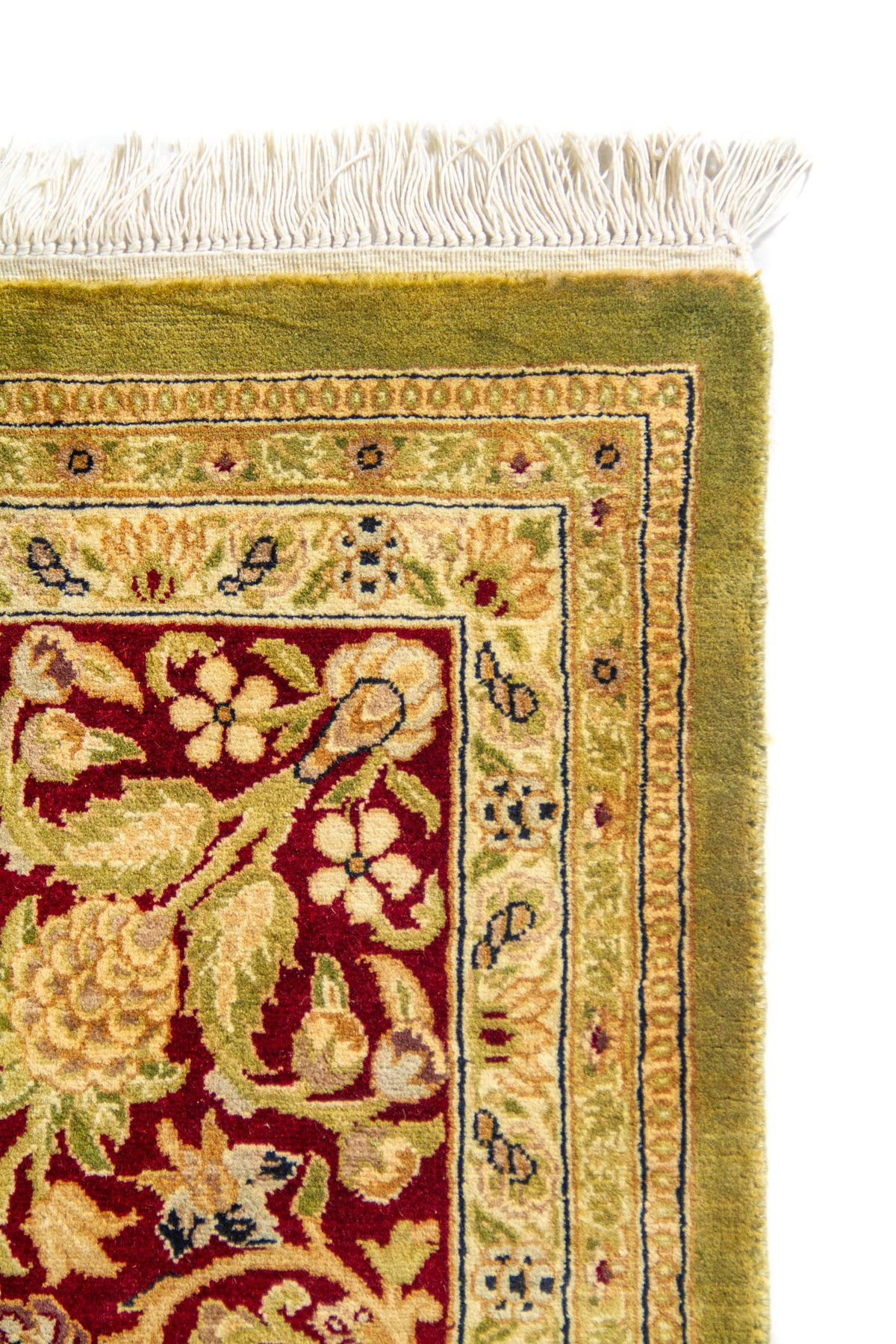 Detalle de esquina superior de alfombra oriental con flecos blancos, borde exterior verde oliva y guarda floral rojo vino con motivos beige.