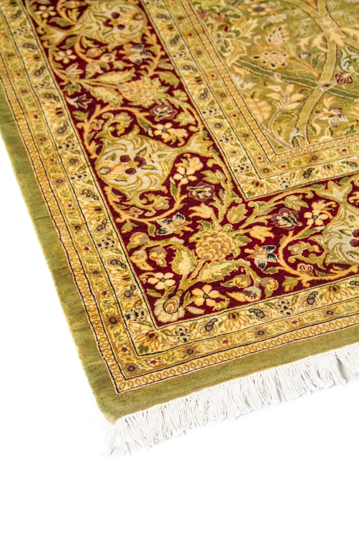 Primer plano de esquina de alfombra oriental en tonos verde, beige y rojo vino, con diseño floral ornamental y acabado con flecos.