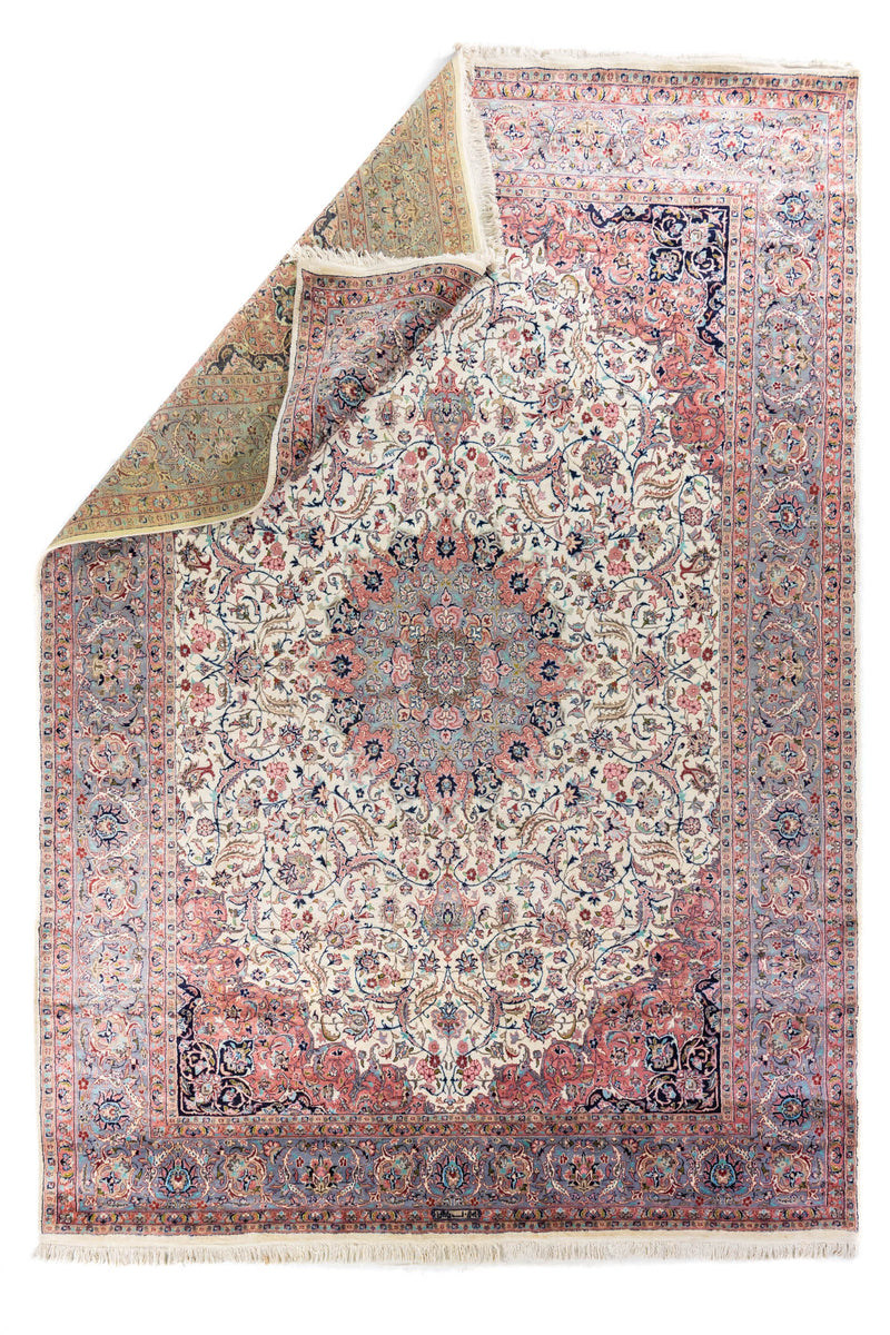 Alfombra persa tradicional doblada mostrando diseño floral central y reverso tejido artesanal con flecos en tono beige.