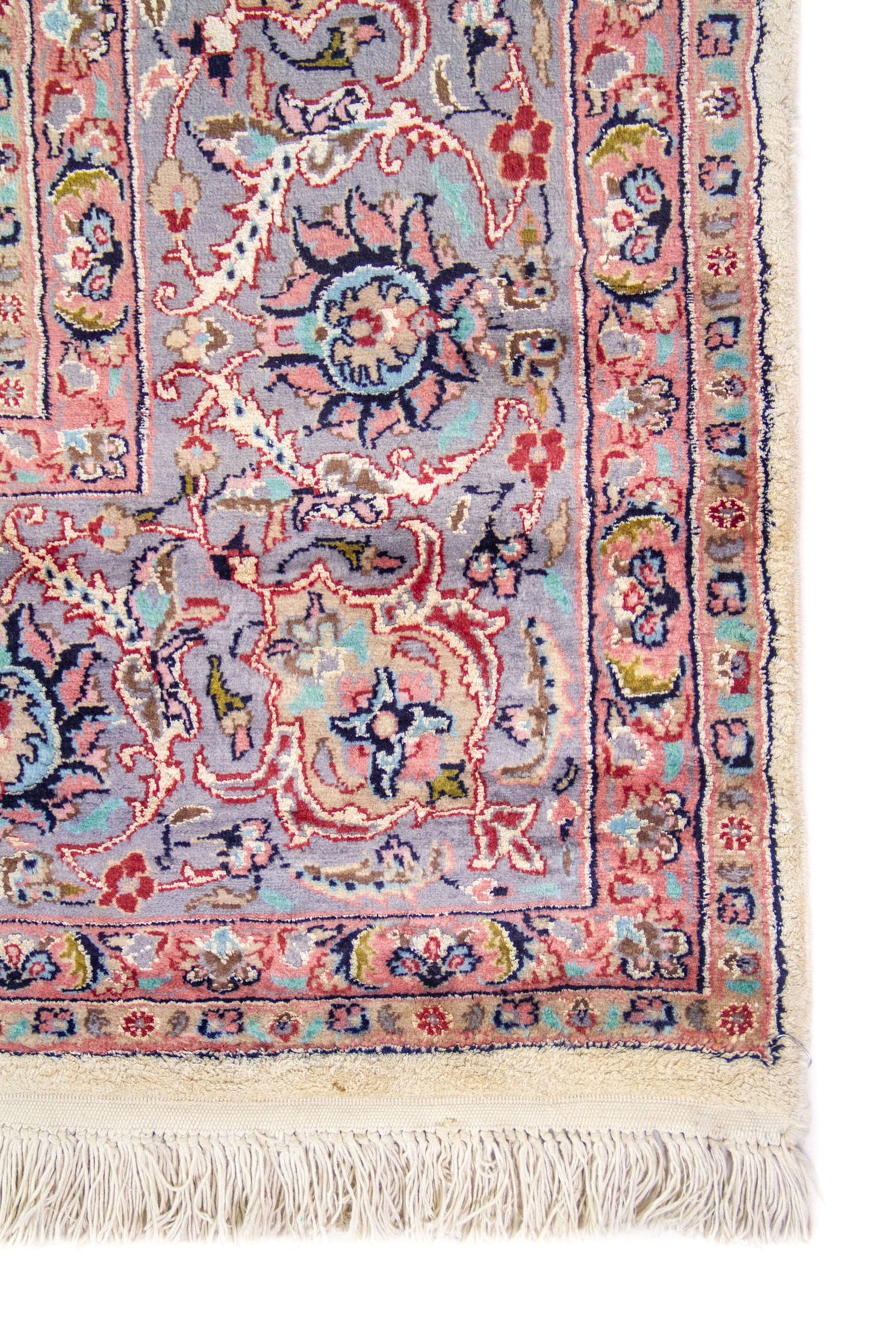 Detalle de esquina de alfombra persa con guarda azul y rosa, motivos florales y flecos tejidos en color beige.