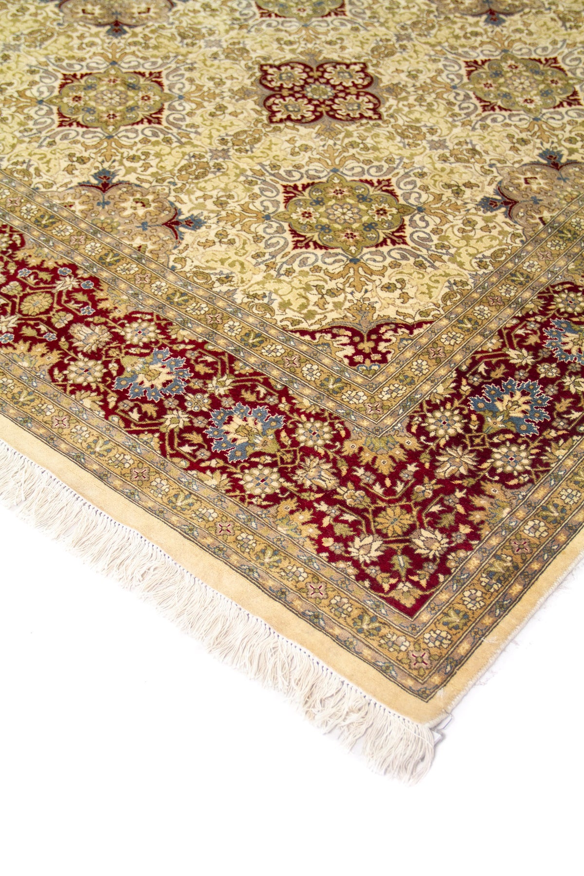 Detalle de esquina de alfombra clásica con campo marfil, motivos florales y guarda roja vino, con flecos color marfil.
