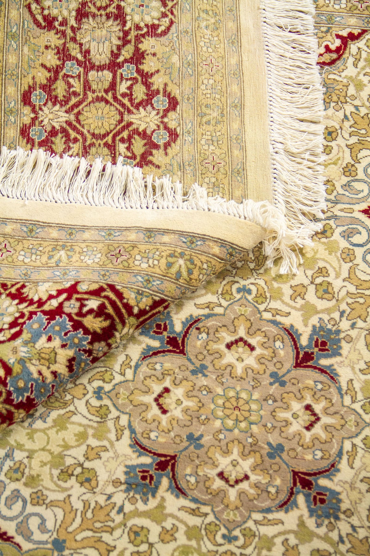 Detalle de alfombra doblada mostrando textura, reverso, flecos y patrón ornamental floral en tonos cálidos.