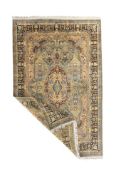 Alfombra ornamental en tonos suaves con una esquina doblada que muestra el reverso y el acabado con flecos