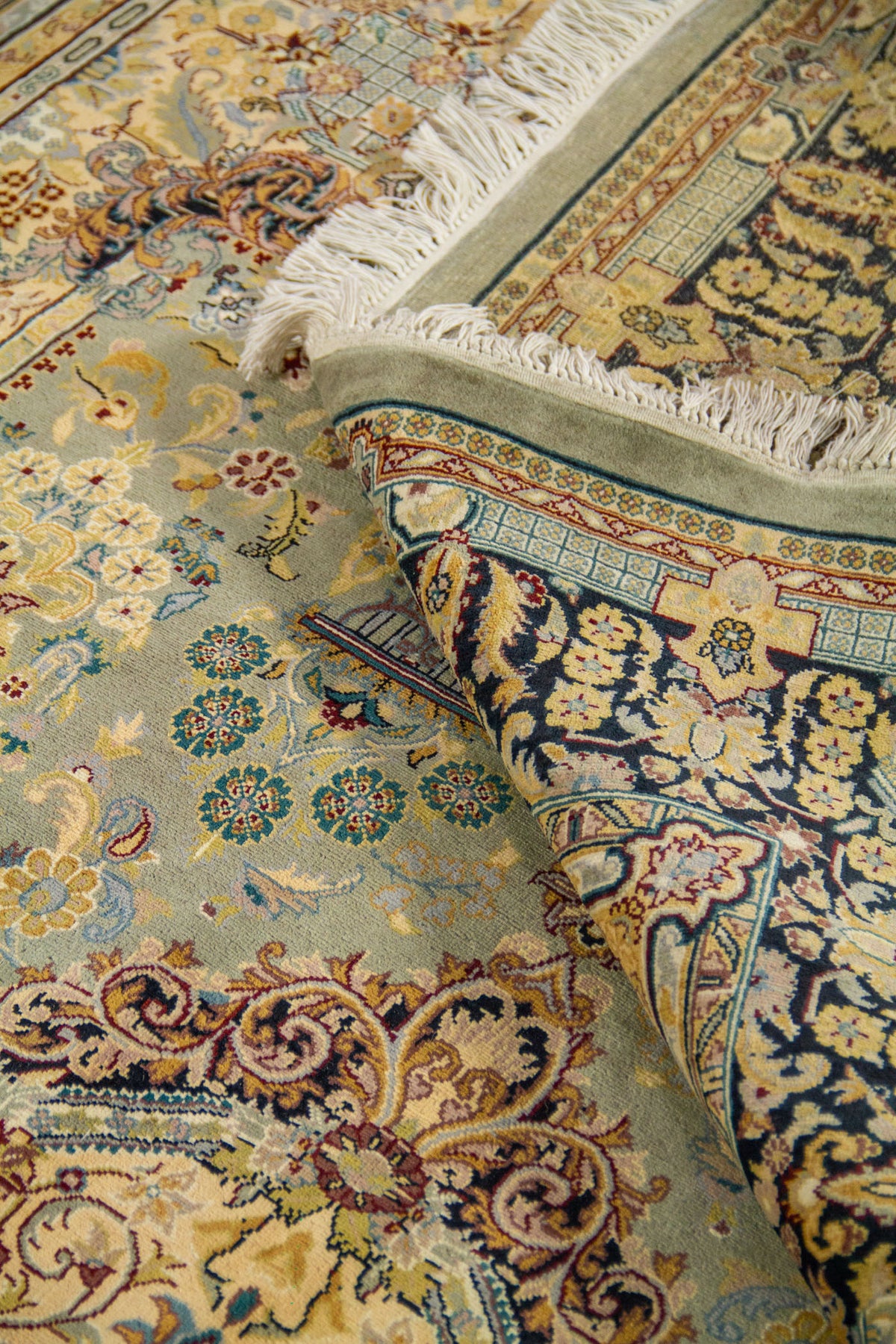 Detalle de alfombra doblada mostrando el reverso, los flecos y los motivos florales en tonos celeste, beige y azul oscuro