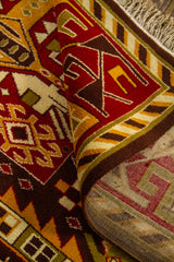 Detalle de alfombra doblada mostrando la textura del tejido, el canto y el reverso, con patrones geométricos en rojo, amarillo y marrón.