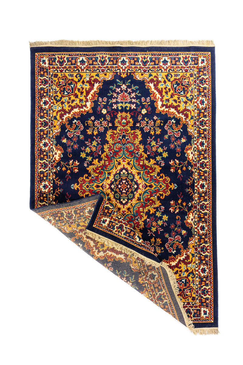 Alfombra oriental azul marino con esquina levantada que muestra su flexibilidad, reverso tejido y terminación con flecos claros.