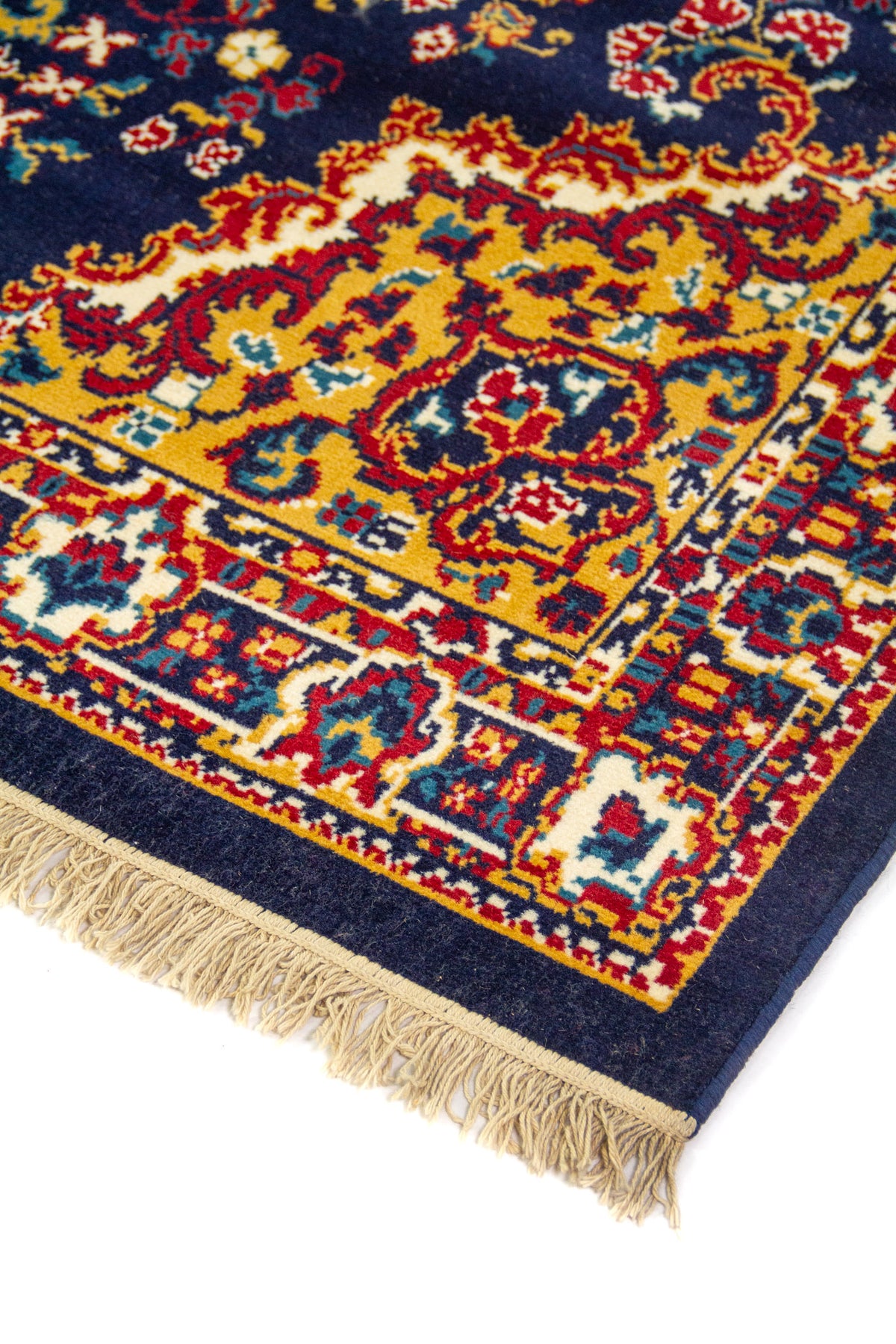 Detalle de esquina de alfombra clásica con fondo azul oscuro, bordes dorados, motivos ornamentales y flecos color beige.