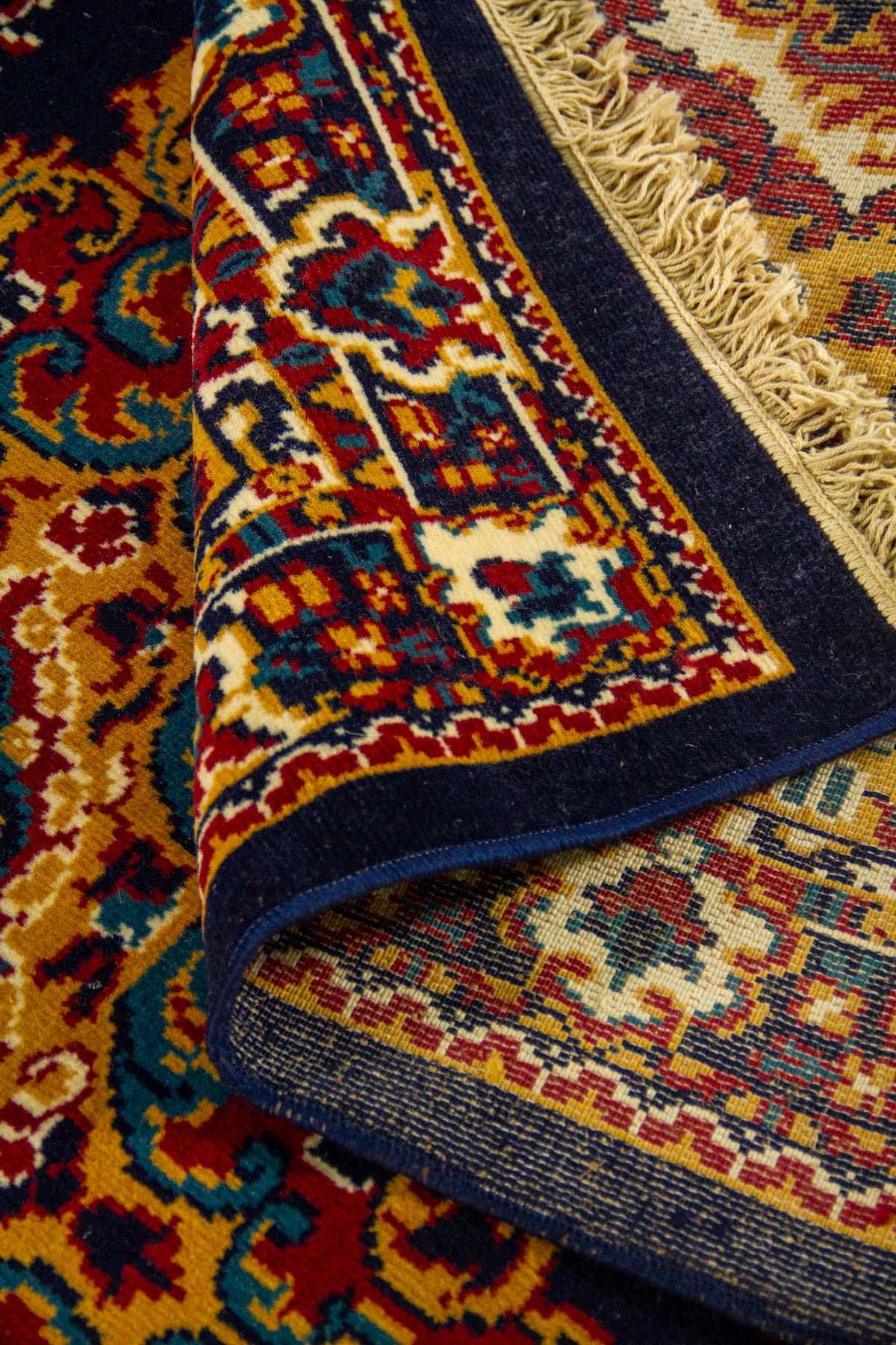 Detalle de alfombra doblada que deja ver el reverso tejido y la riqueza del diseño ornamental en tonos azul, dorado y rojo.