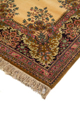 Detalle en perspectiva de alfombra clásica con fondo beige cálido, borde floral oscuro y terminación con flecos claros.