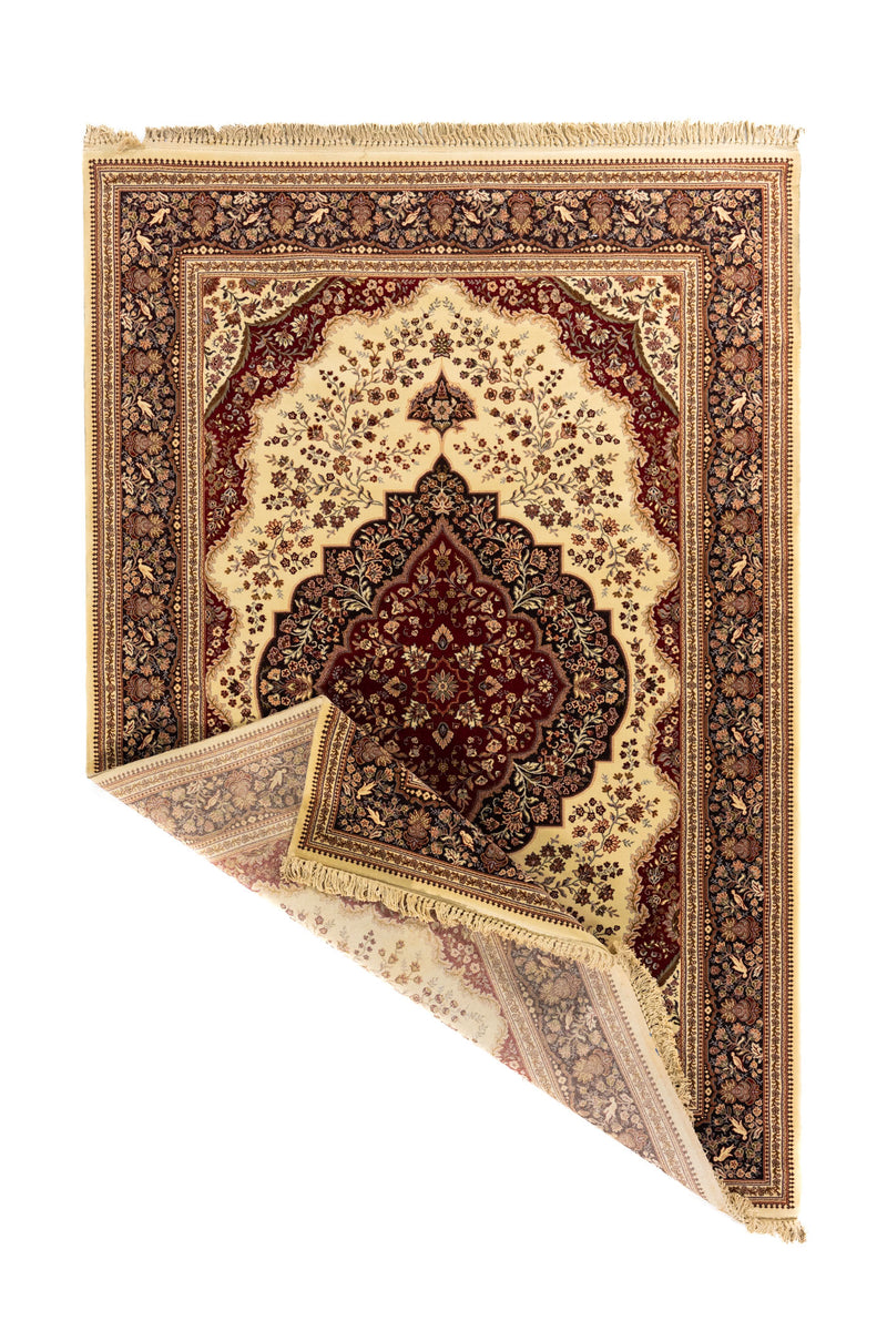 Alfombra oriental en tonos beige, rojo y marrón oscuro con una esquina doblada que deja ver el reverso y el acabado con flecos.