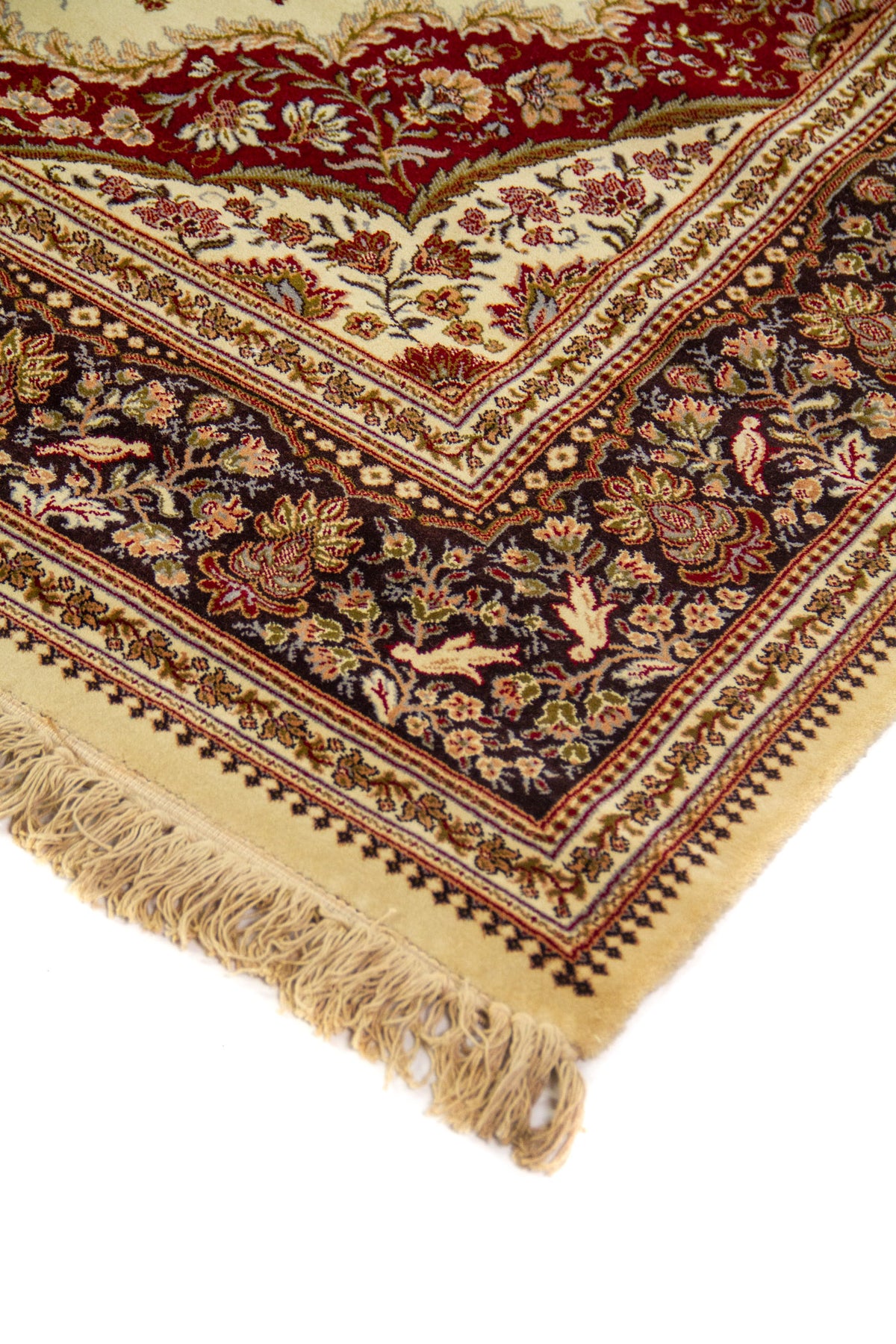 Detalle de esquina de alfombra clásica con motivos florales, fondo beige, borde oscuro ornamental y flecos color beige.