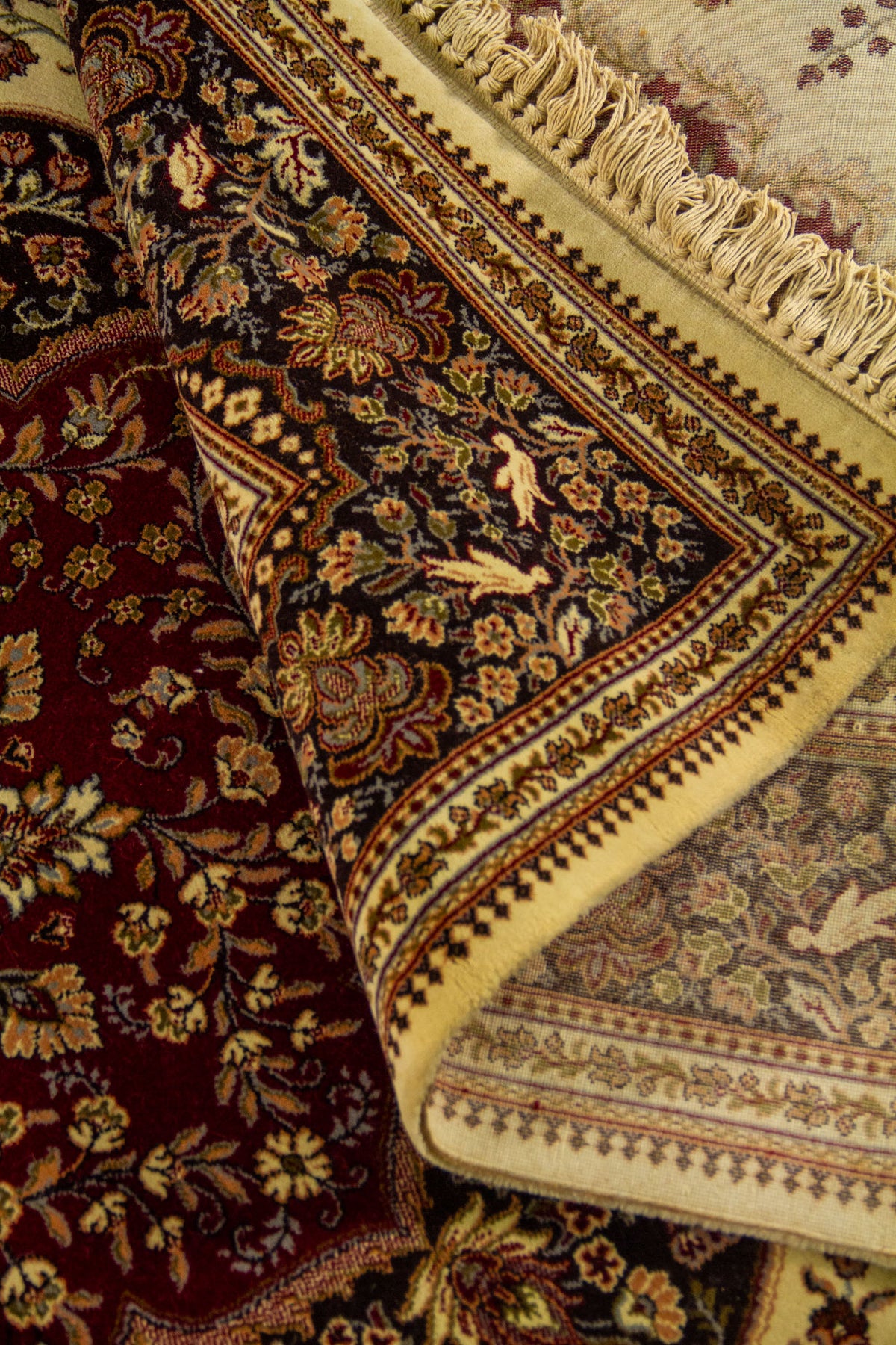 Detalle de alfombra doblada mostrando el reverso, los flecos y los motivos florales en tonos beige, rojo, marrón y dorado.