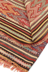 Alfombra kilim con diseño de rayas y patrones geométricos en tonos rojos, verdes y beige, mostrando detalles intrincados y flecos.