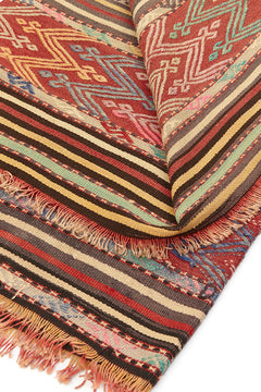 Alfombra kilim con diseño de rayas y patrones geométricos en tonos rojos, verdes y beige, mostrando detalles intrincados y flecos.