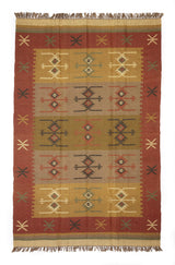 Alfombra de estilo kilim con patrones geométricos en tonos cálidos y detalles de flecos, parcialmente doblada mostrando su reverso.