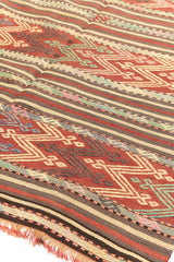 Alfombra kilim con diseño de rayas y patrones geométricos en tonos rojos, verdes y beige, mostrando detalles intrincados y flecos.