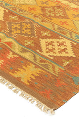 Alfombra kilim con diseño de figuras geométricas en tonos dorados, marrones y naranjas, con borde de flecos decorativos.