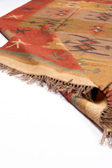 Alfombra de estilo kilim con patrones geométricos en tonos cálidos y detalles de flecos, parcialmente doblada mostrando su reverso.