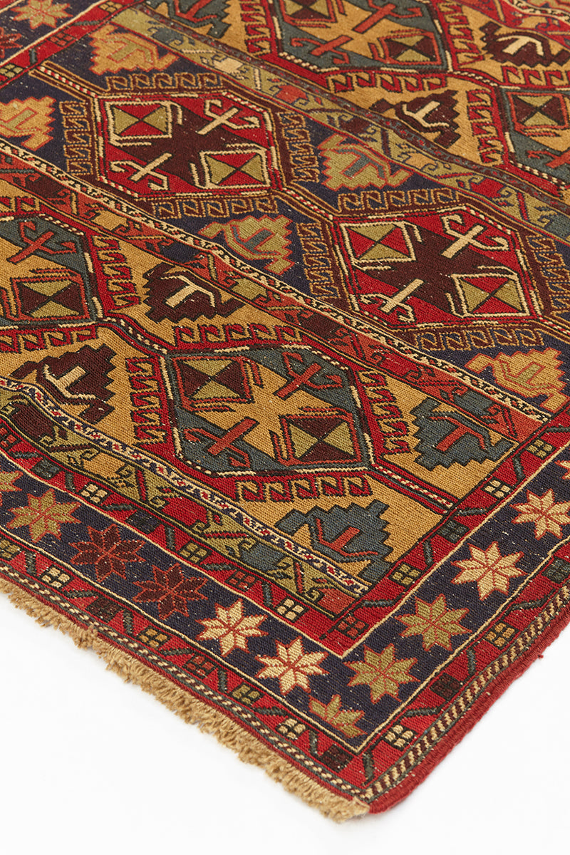 Alfombra kilim con diseño de rombos y figuras geométricas en tonos rojos, dorados y azul marino, con borde de flecos y patrones intrincados.