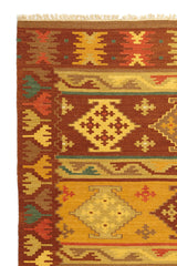 Alfombra kilim con motivos geométricos en tonos marrones, dorados y naranjas, con borde de flecos y detalles intrincados.