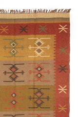 Alfombra de estilo kilim con patrones geométricos en tonos cálidos y detalles de flecos, parcialmente doblada mostrando su reverso.