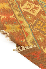 Alfombra kilim con diseño de figuras geométricas en tonos dorados, marrones y naranjas, con borde de flecos decorativos.